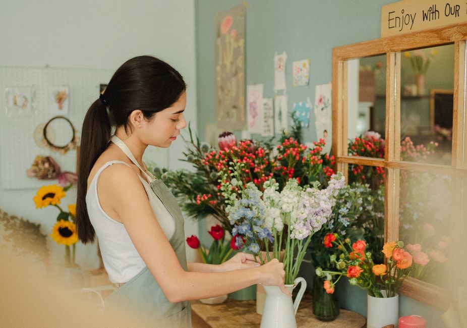 A young woman with dark hair tied back, dressed in a white sleeveless top and a light green apron, is arranging a bouquet of fresh flowers inside a florist shop. She is placing light purple and white 