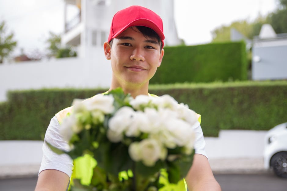 A delivery person wearing a red cap and a high-visibility vest is smiling and extending a bouquet of fresh white flowers towards the camera. The bouquet features tightly arranged blooms with green fol