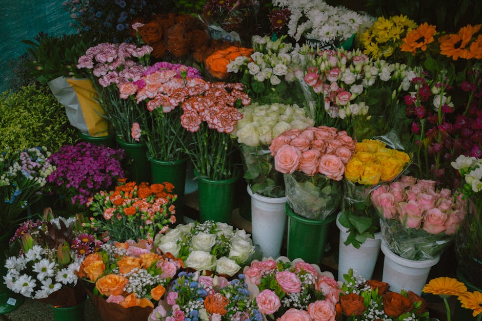 A vibrant display of fresh flowers arranged in various bouquets and potted plants, including pink, white, yellow, orange, purple, and red blooms. The flowers are wrapped in clear or colored plastics, 