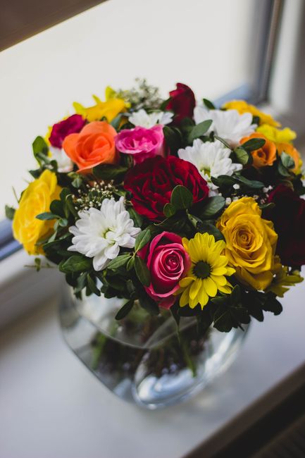 A vibrant floral bouquet arranged in a clear glass vase, featuring a mix of fresh roses in shades of red, pink, yellow, and orange, complemented by white daisies, yellow sunflowers, and greenery. The 
