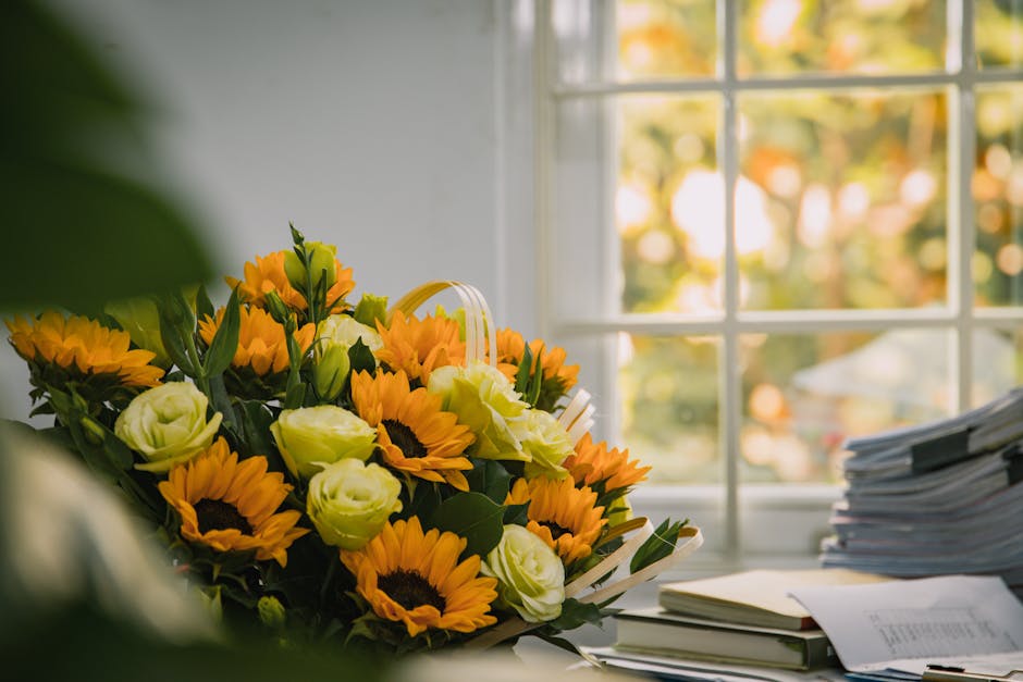A vibrant floral arrangement featuring fresh sunflowers with bright yellow petals and dark brown centers, complemented by white roses and small yellow blossoms, set on a desk near a window. The bouque