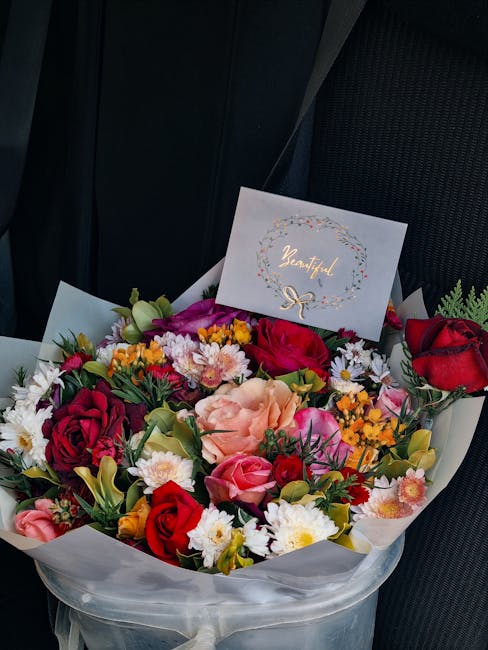 A vibrant floral arrangement featuring a mix of roses, carnations, chrysanthemums, and small yellow and white filler flowers, with colours including deep red, soft pink, peach, purple, and white. The 