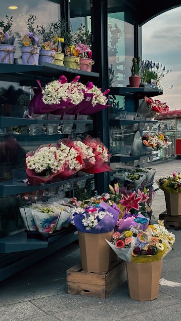 A display of fresh floral bouquets arranged on metal shelves in a flower shop, featuring a variety of colorful flowers including white chrysanthemums, pink lilies, and purple lisianthus, wrapped in pa